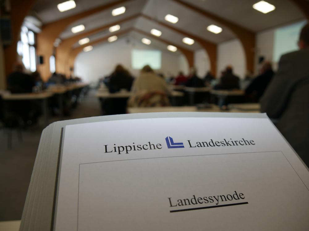 Im Vordergrund ist eine Mappe zu sehen. Darauf steht: Lippische Landeskirche und dadrunter  Landessynode. Im Hintergrund sieht man verschwommen Personen, die in einem großen Raum sitzen. 