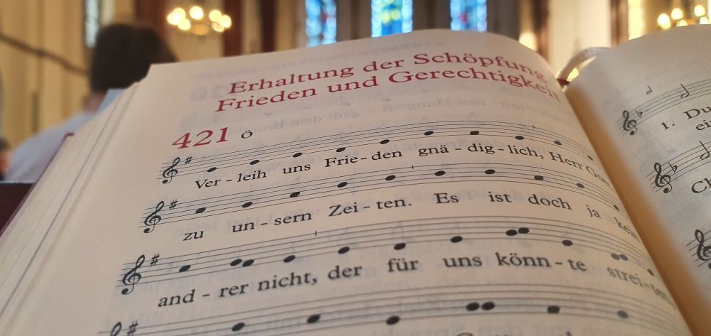 . Im Vordergrund ein aufgeschlagenes Gesangbuch. Zu sehen sind die ersten Zeilen des Liedes Nr. 421 „Verleih und Frieden, gnädiglich“ . Oben drüber steht der Themenkreis : Erhaltung der Schöpfung. Frieden und Gerechtigkeit. Im Hintergrund sind schemenhaft die bunten Glasfenster einer Kirche zu erkennen. 